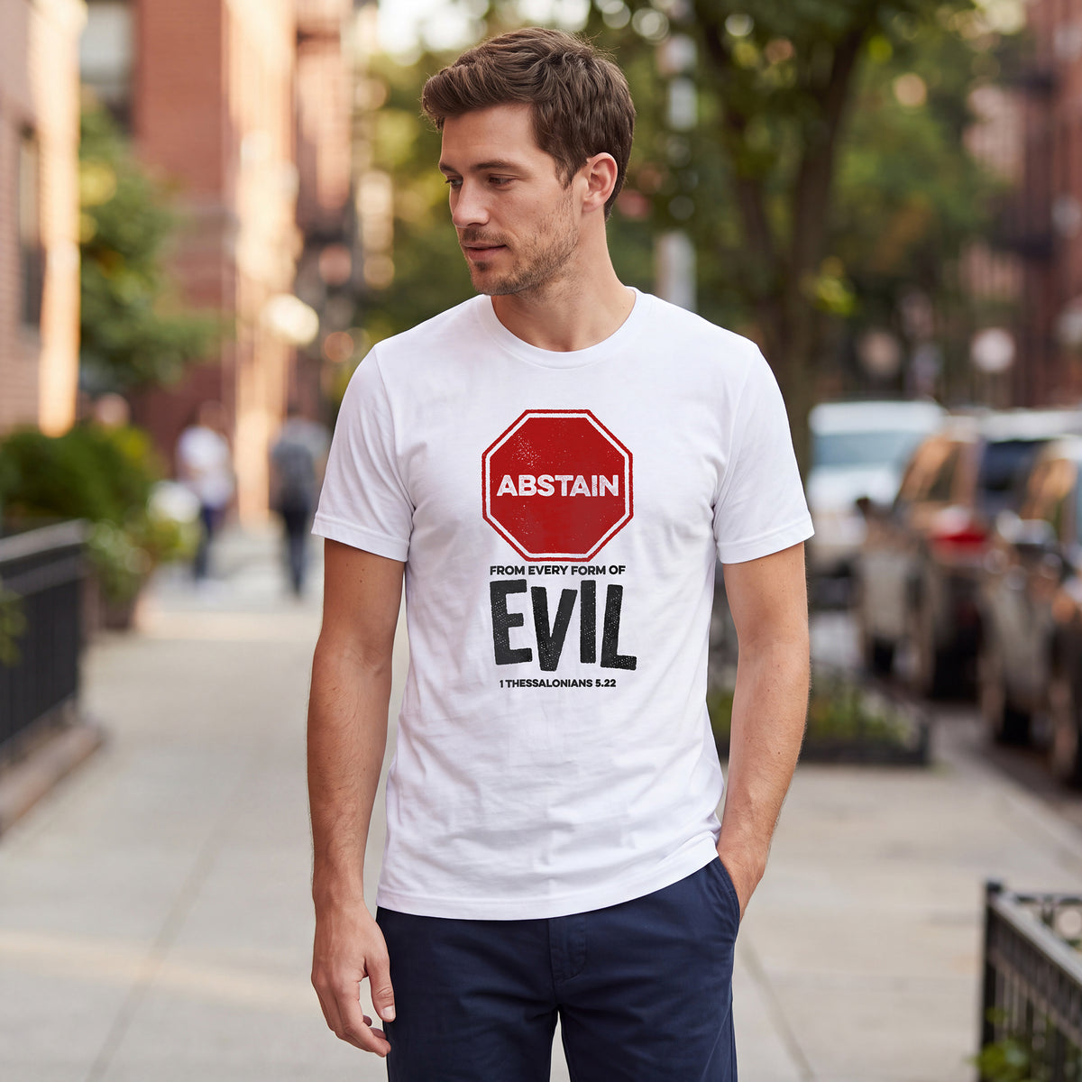Man standing on an urban street wearing a white t-shirt with 1 Thessalonians 5.22 graphic printed on the front.