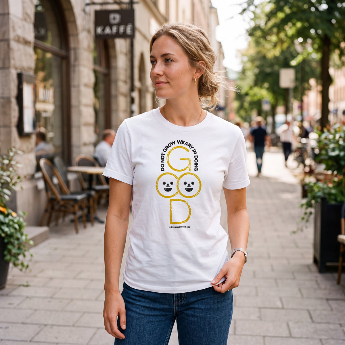 Woman on city sidewalk wearing white t-shirt with 2 Thessalonians 3:13 graphic print.