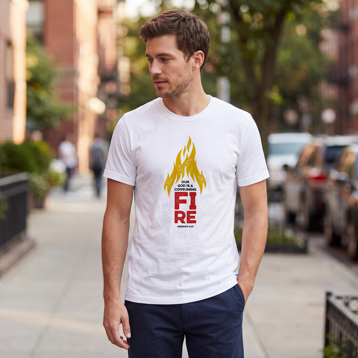 Man standing on an urban street wearing a white t-shirt with Hebrews 12.29 graphic printed on the front.